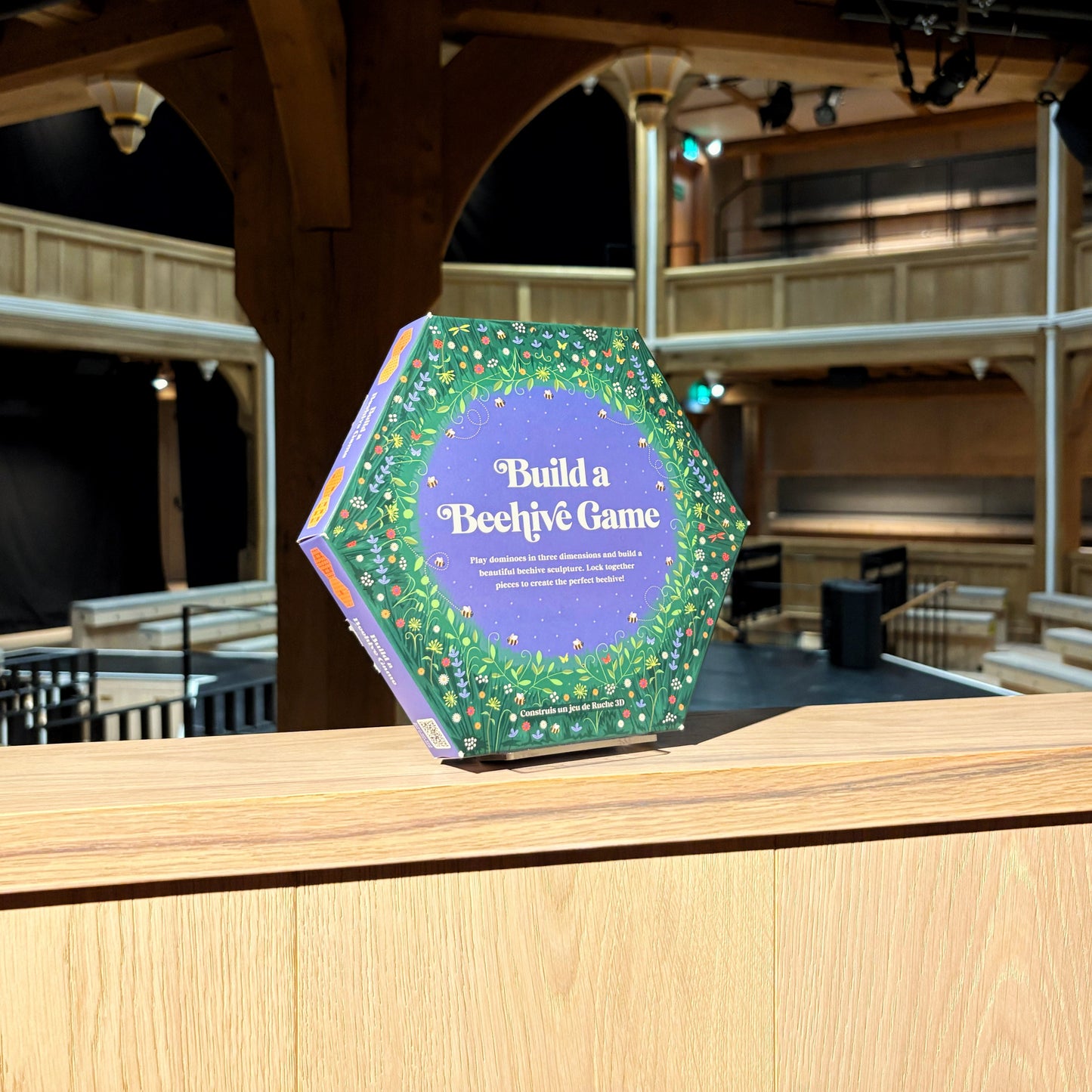 Hexagonal game box titled 'Build a Bechive Game' on a wooden platform with theater seats in the background