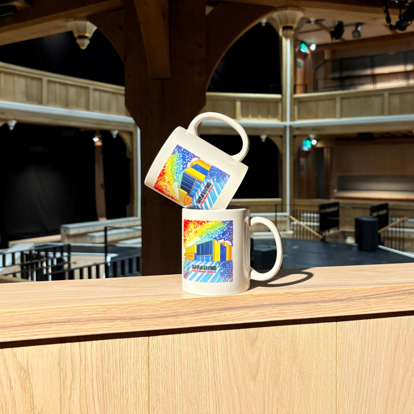 Two mugs stacked, each features a rainbow illustration of the Shakespeare North building