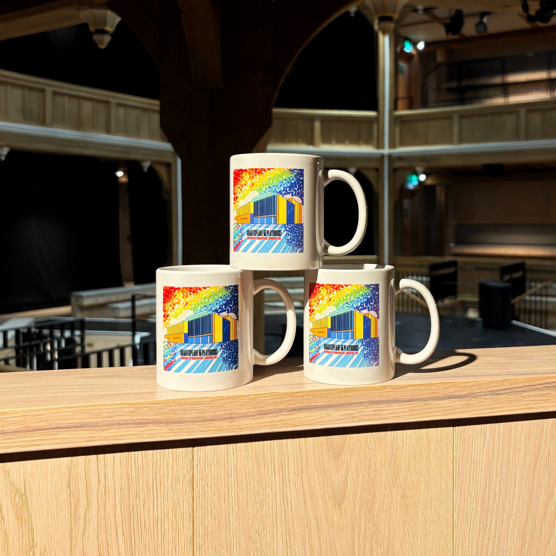 Three mugs stacked, each featuring a rainbow illustration of the front of the Shakespeare North building.